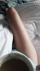 Woman sitting in bed drinking tea looking down her body to her legs