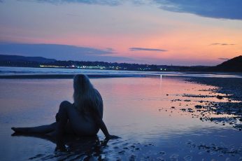 woman sitting naked on the shore with dusty pink sunset behind her