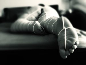Woman laying face down on bed bound in white rope with it passing between her toes