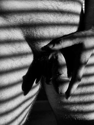 black and white of man holding his penis with shadow lines from a blind running over his body