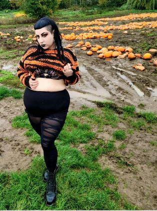 Devvie wearing black shirt and halloween stripped jumper holding her top up showing her bra in pumpkin patch