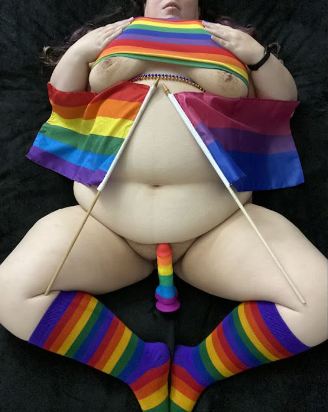 Ash laying on her back wearing pride socks, bra, flags with a pride dildo between her legs