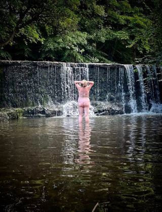 Naked woman standing in river with back to the camera and waterfall in front of her