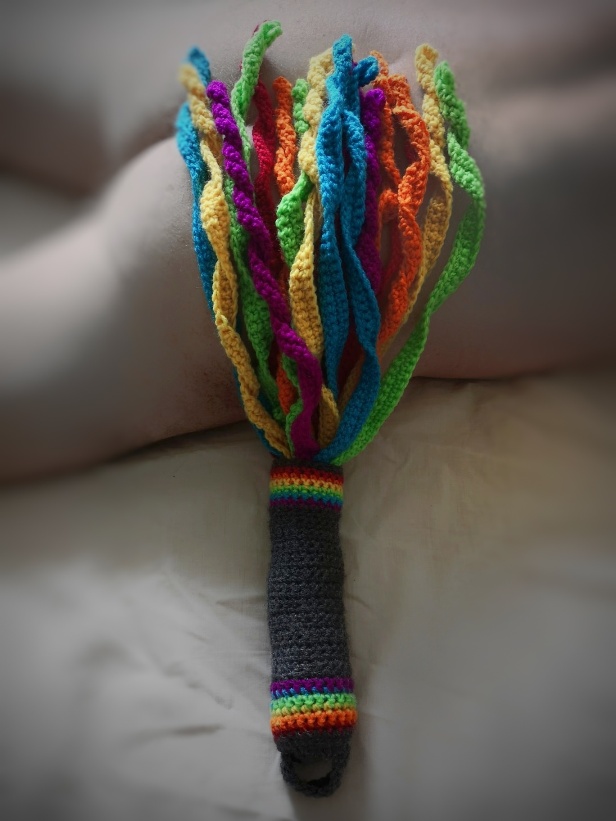 Man laying naked face down on bed with rainbow colours flogger laying on one butt cheek
