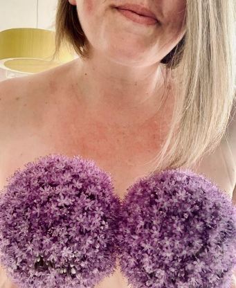 Woman topless with purple alliums like pompoms in front of her breasts