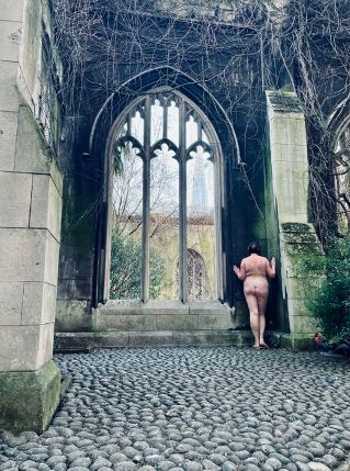 Woman naked standing in the corner of ruined church with the Shard london building just visible through the window frames