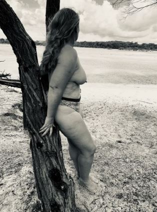 Black and white of woman topless standing on the shore leaning against drift wood