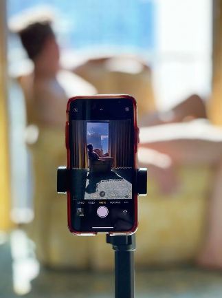 Phone on tripod. On the screen is the picture in the distance which is someone sitting naked on a chair in front of a large window. In the background we can see the larger blurred image that is inside the screen