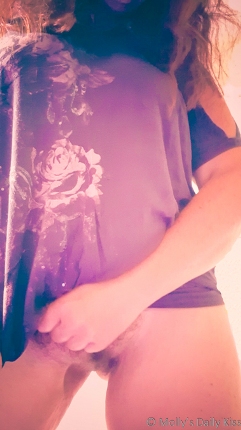 Molly standing wearing a pinky grey t shirt and nothing else. Her had is grabbing her pubes