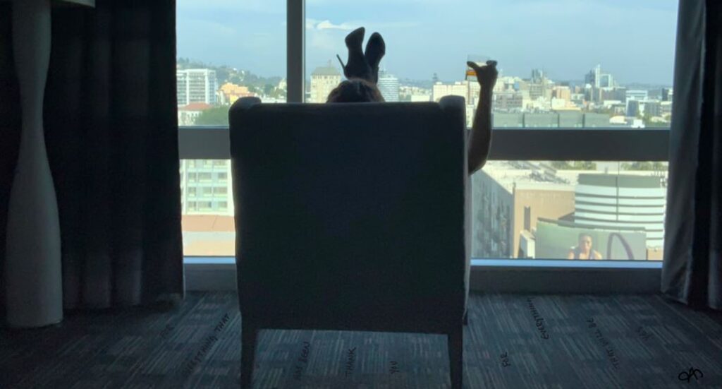 Cropped image by Annie Savoy of woman in high heels sitting in chair in front of large window looking out over the city