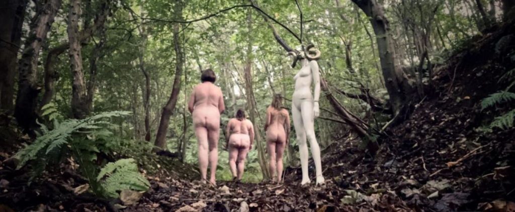 Three nude women with backs to the camera in woodland setting with a mannequin with a ram head behind them
