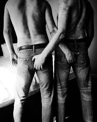 Two men wearing jeans and topless standing with their back to the camera and reaching to grab each others butt