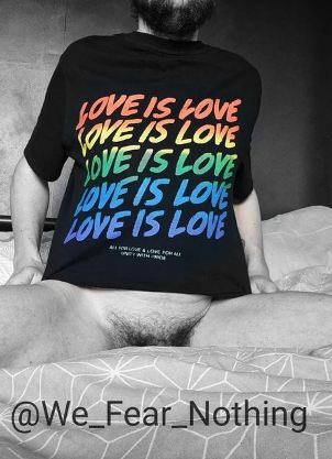 Person with beard sitting on bed with legs open. We can see the top of their pubic hair and they are wearing a black t shirt with the words love is love in pride colours 
