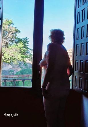 Nude woman standing in front of large windows with wooden shutters looking out at a summer day. 