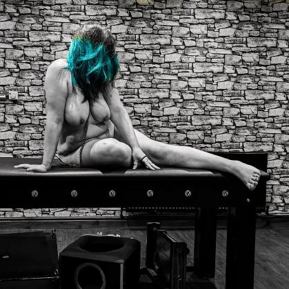 Black and white of nude woman sitting on high u leather bench with brick wall behind her. The only colour in the image is her blue hair