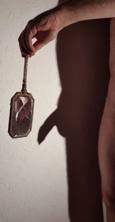 Nude man casting shadow on the wall. He is holding a small hand mirror in front of himself and you can see his penis reflected in the mirror
