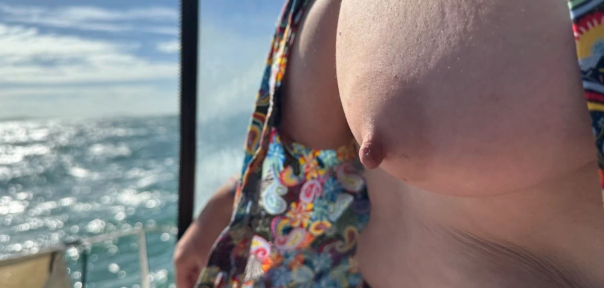 Person with the front of their wrap dress pulled up to show their naked breasts with the sea shimming in the sunlight beyond them
