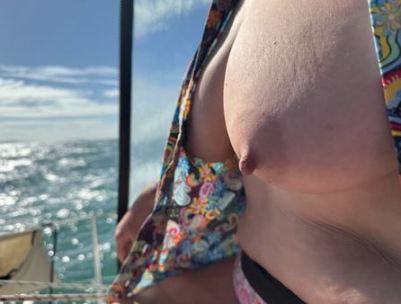 Person with the front of their wrap dress pulled up to show their naked breasts with the sea shimming in the sunlight beyond them