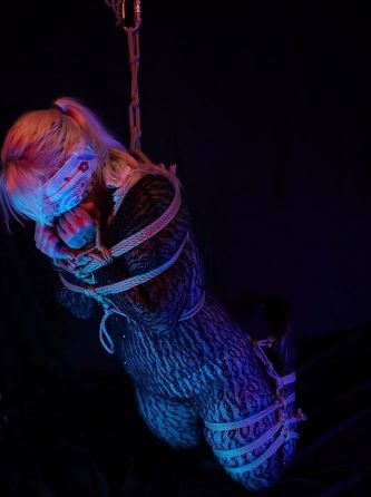 Bare foot sub in rope suspension. The back ground is black and she is lit by a purple pink light. Her face is covered with a scarf for weekly round-up 701