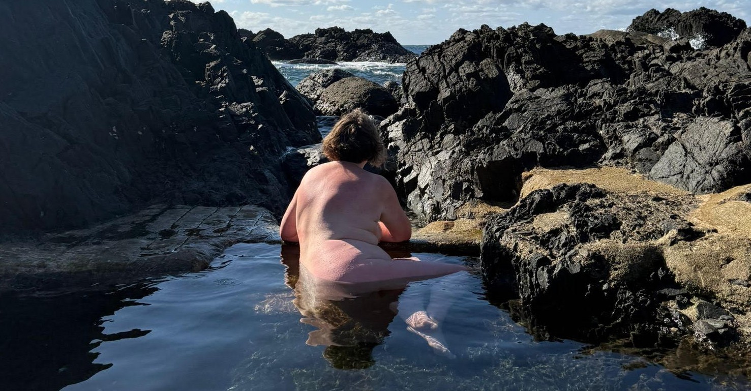 Cropped image of Exposing 40 sitting in rockpool naked arrounded by black rocks. The water is crystal clear and the sky is blue for post called Day 3653