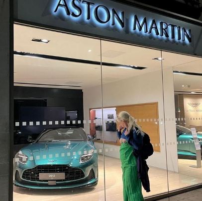 Barefoot sub in green dress standing in front of Aston Marton car dealership with her boobs showing